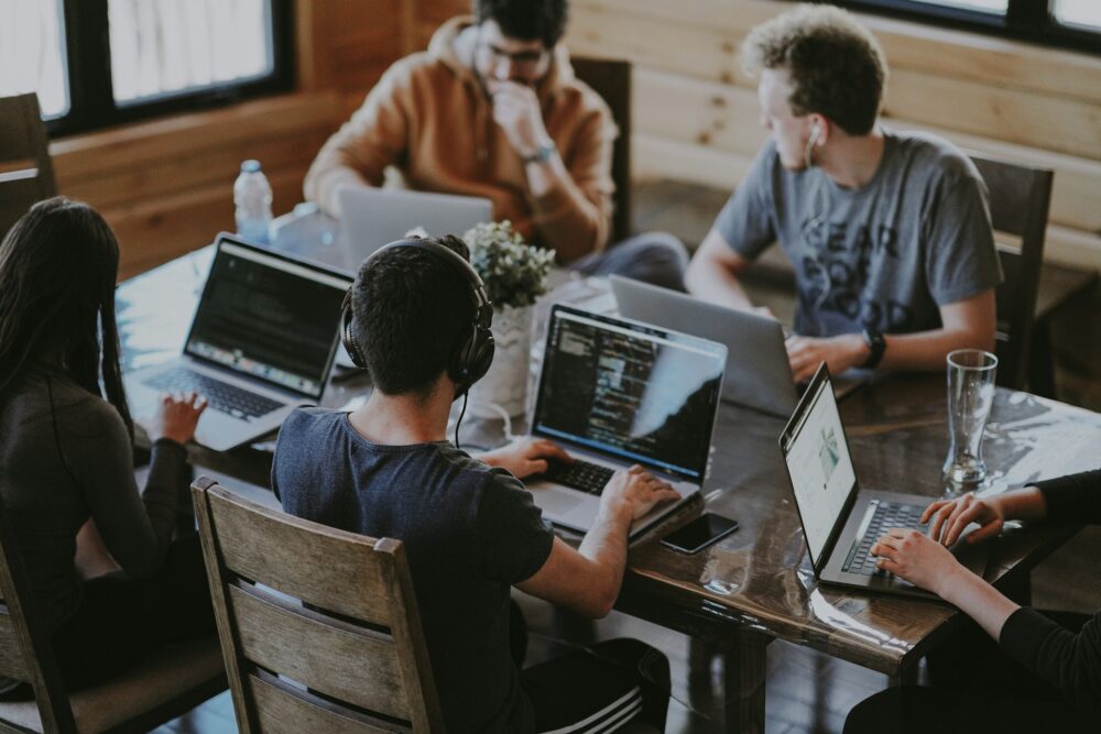 Five coworkers work on laptops around a shared wooden table, one in headphones, showing flexible workplace strategies at work