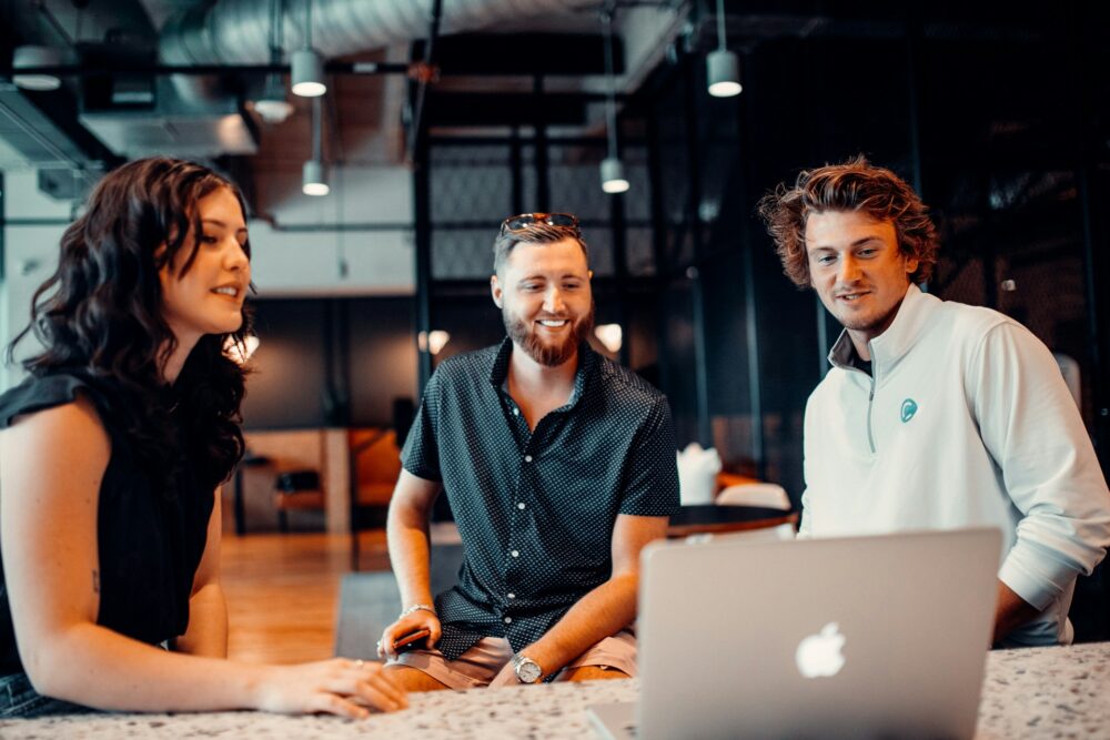 Three coworkers smile while reviewing a laptop together in an open industrial office, an example of workplace flexibility