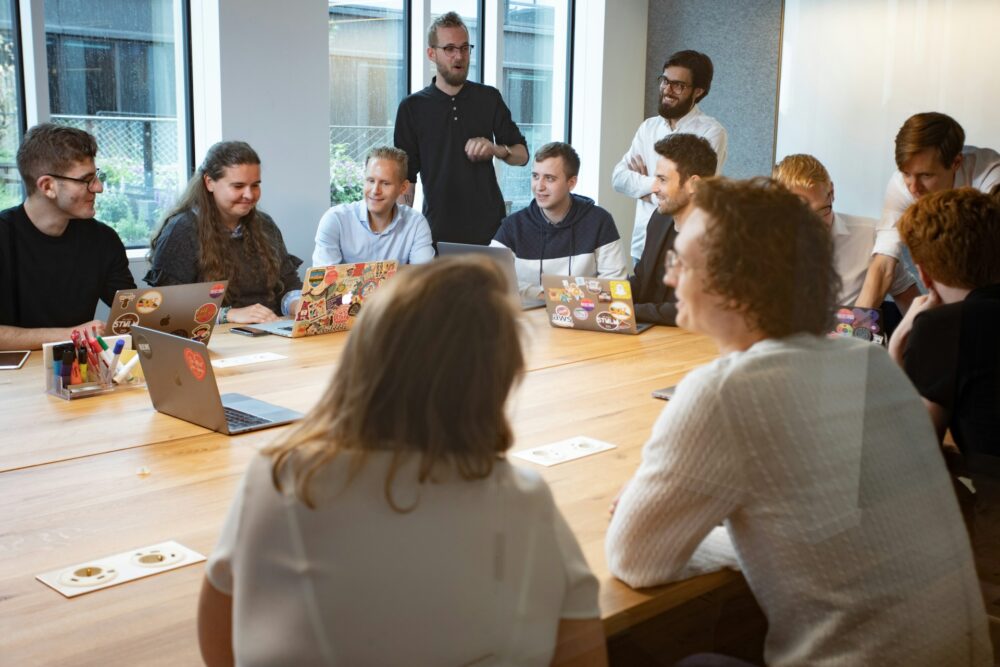 Team members around a long conference table with laptops laugh as a colleague speaks, reflecting a flexible work culture
