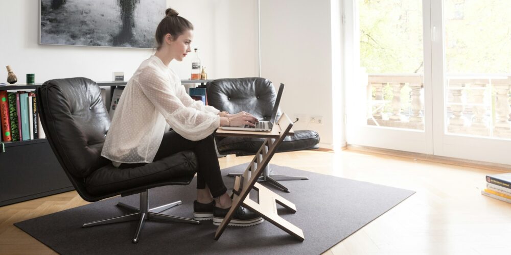 Woman types on a laptop at a wooden standing desk beside a lounge chair in a bright home office, a flexible work environment