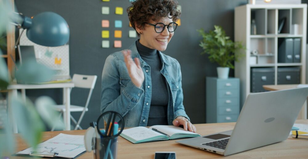 Smiling woman in glasses waves at her laptop during a video call from her office desk, illustrating a flexible workplace