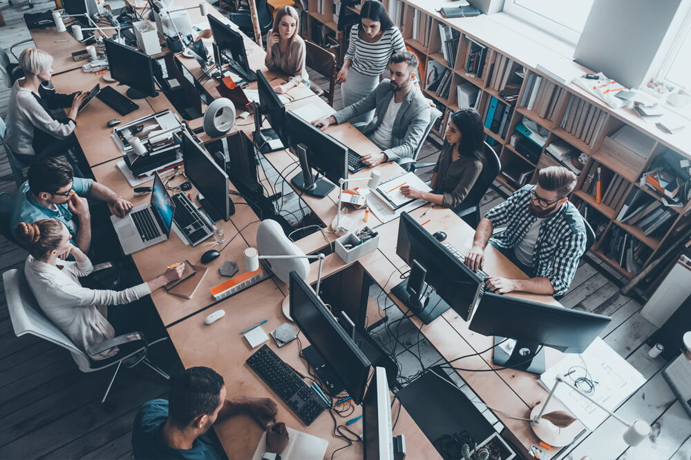 Overhead view of a busy open-plan office. Visualizing the massive scale of the corporate workforce and overall ai impact on jobs.