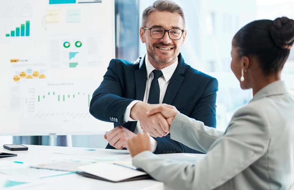 Finance director recruitment handshake between two executives in front of a whiteboard filled with marketing and revenue projection charts